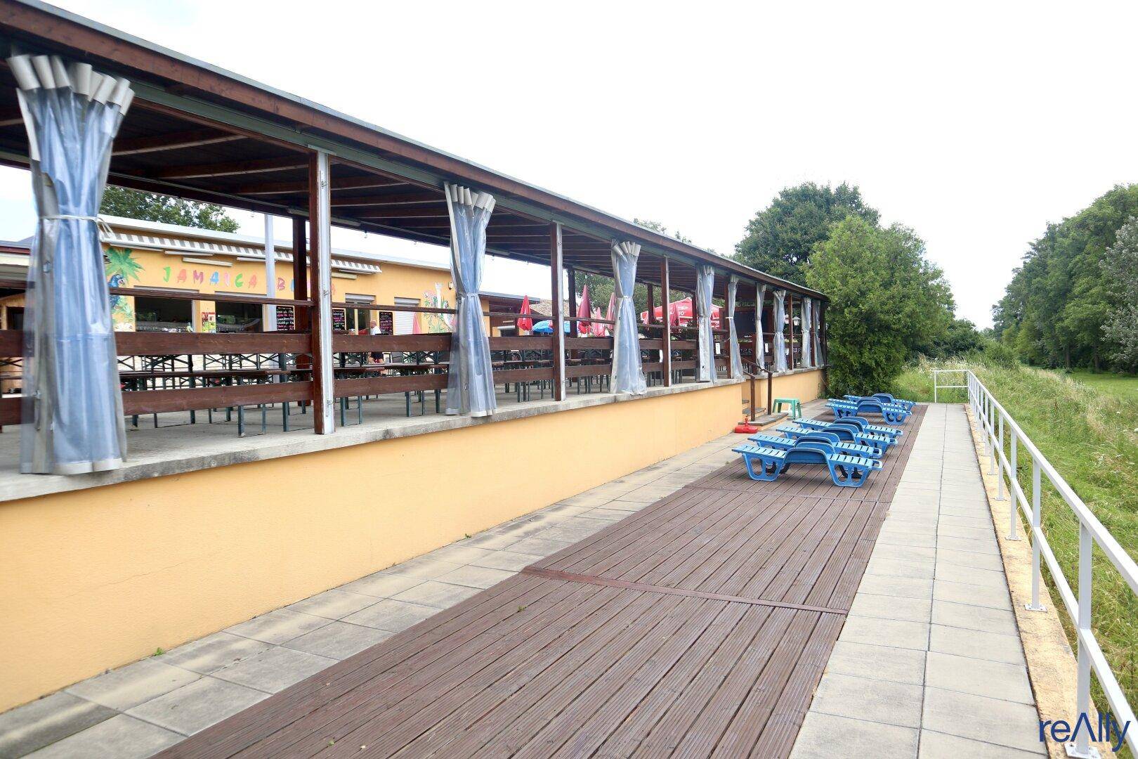 Überdachter Gang oder Terrasse mit Holzbelag und blauen Liegestühlen, mit Blick auf die Umgebung.