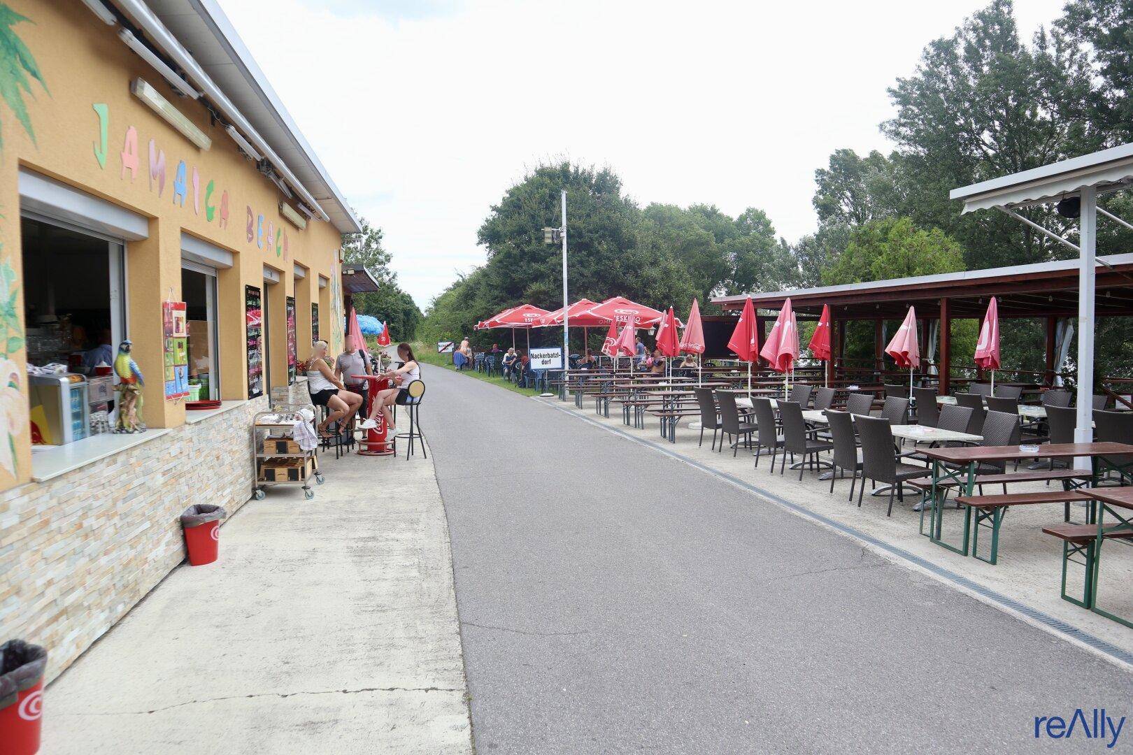 Langer Außenbereich eines Restaurants mit vielen Tischen, Stühlen und roten Sonnenschirmen unter Bäumen.