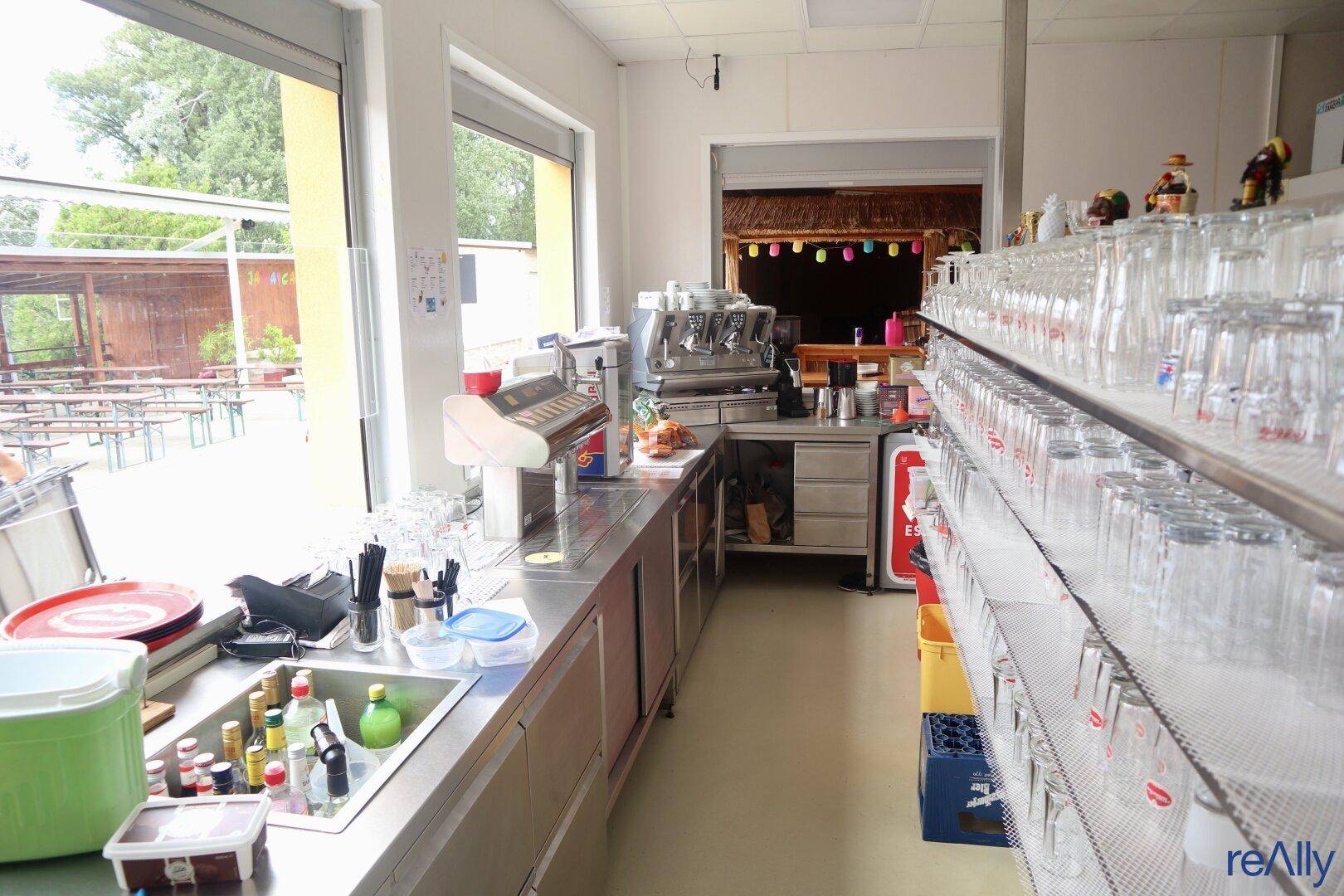 Innenansicht einer Bar mit Edelstahltheke, Kaffeemaschine und vielen Gläsern, mit Blick nach draußen.