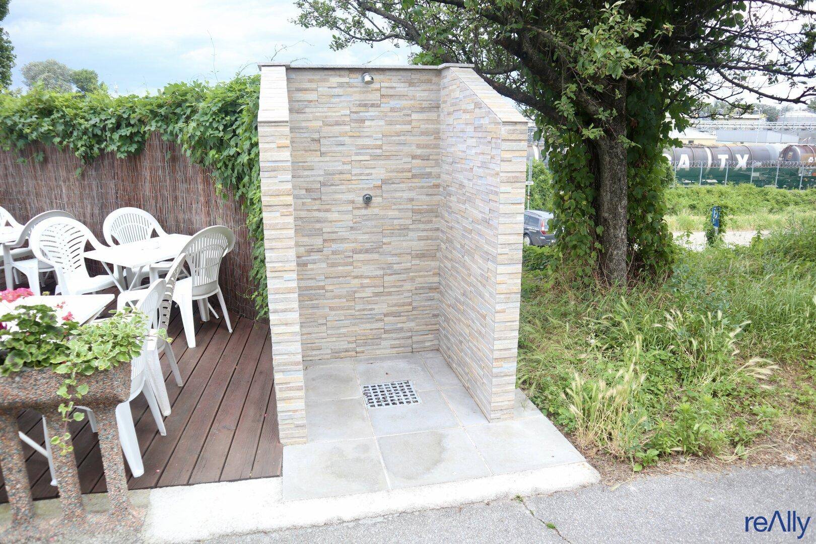 Eine Außendusche mit Steinverkleidung, umgeben von Grünflächen und einer Holzterrasse.
