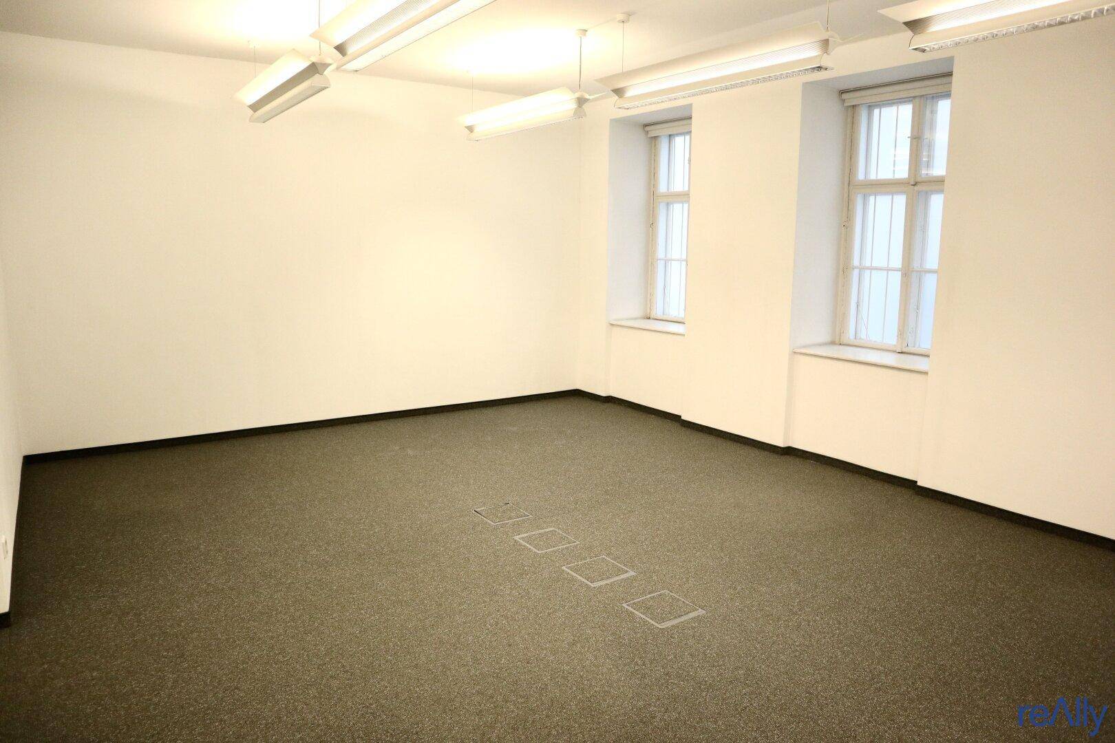 Heller Büroraum mit Teppichboden, großen Fenstern und Deckenleuchten, ideal für Arbeitsbereiche.