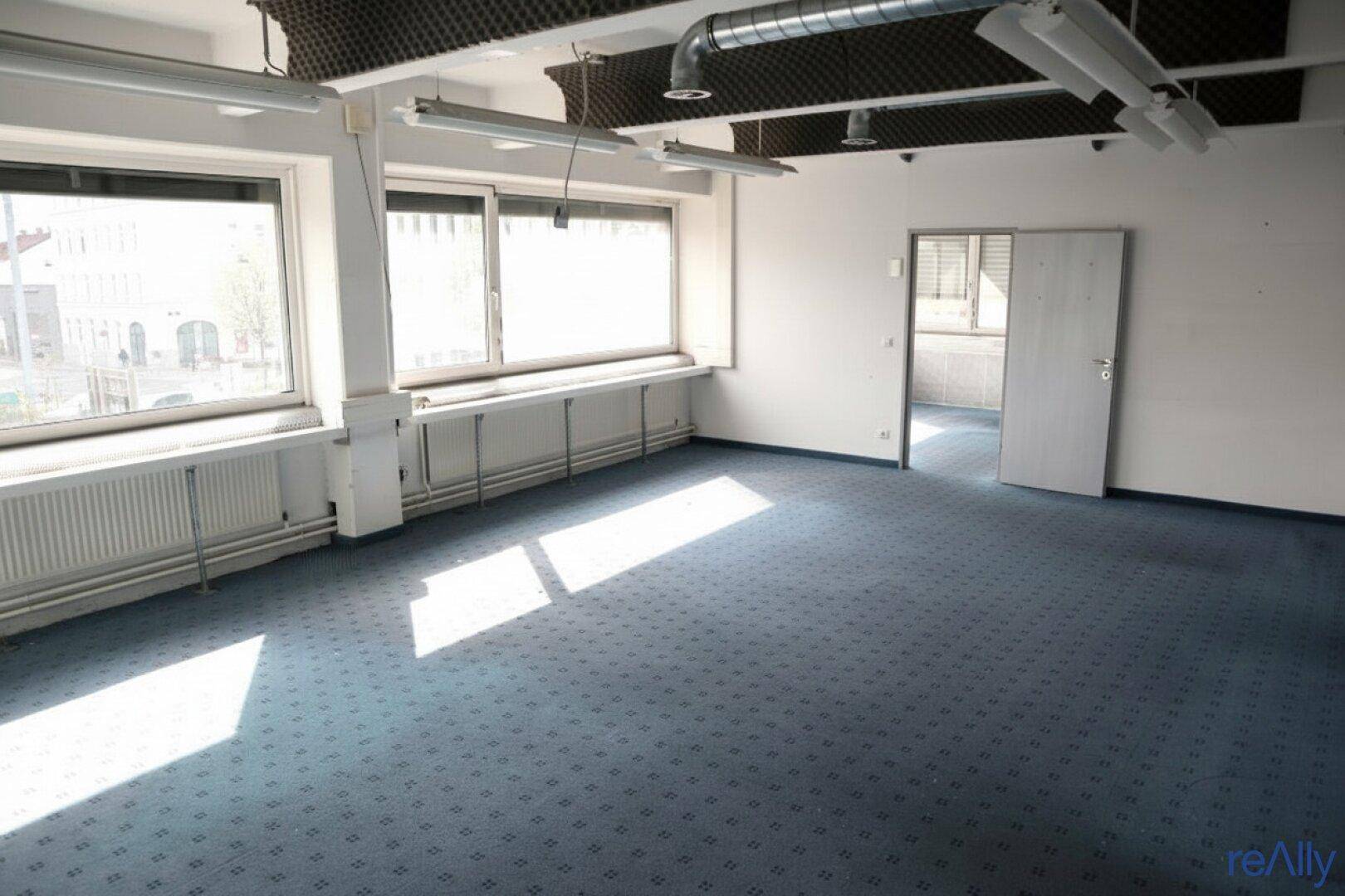Großes Büro mit blauem Teppichboden, mehreren Fenstern und sichtbaren Deckeninstallationen.