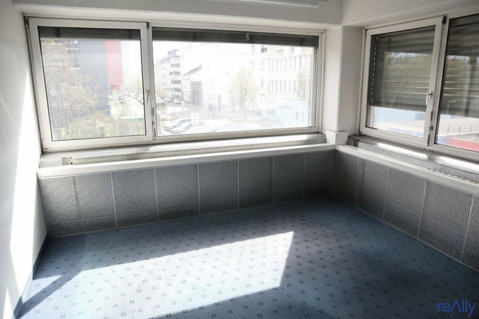 Eckbüro mit blauem Teppichboden, großen Fenstern und viel natürlichem Licht, das den Raum erhellt.