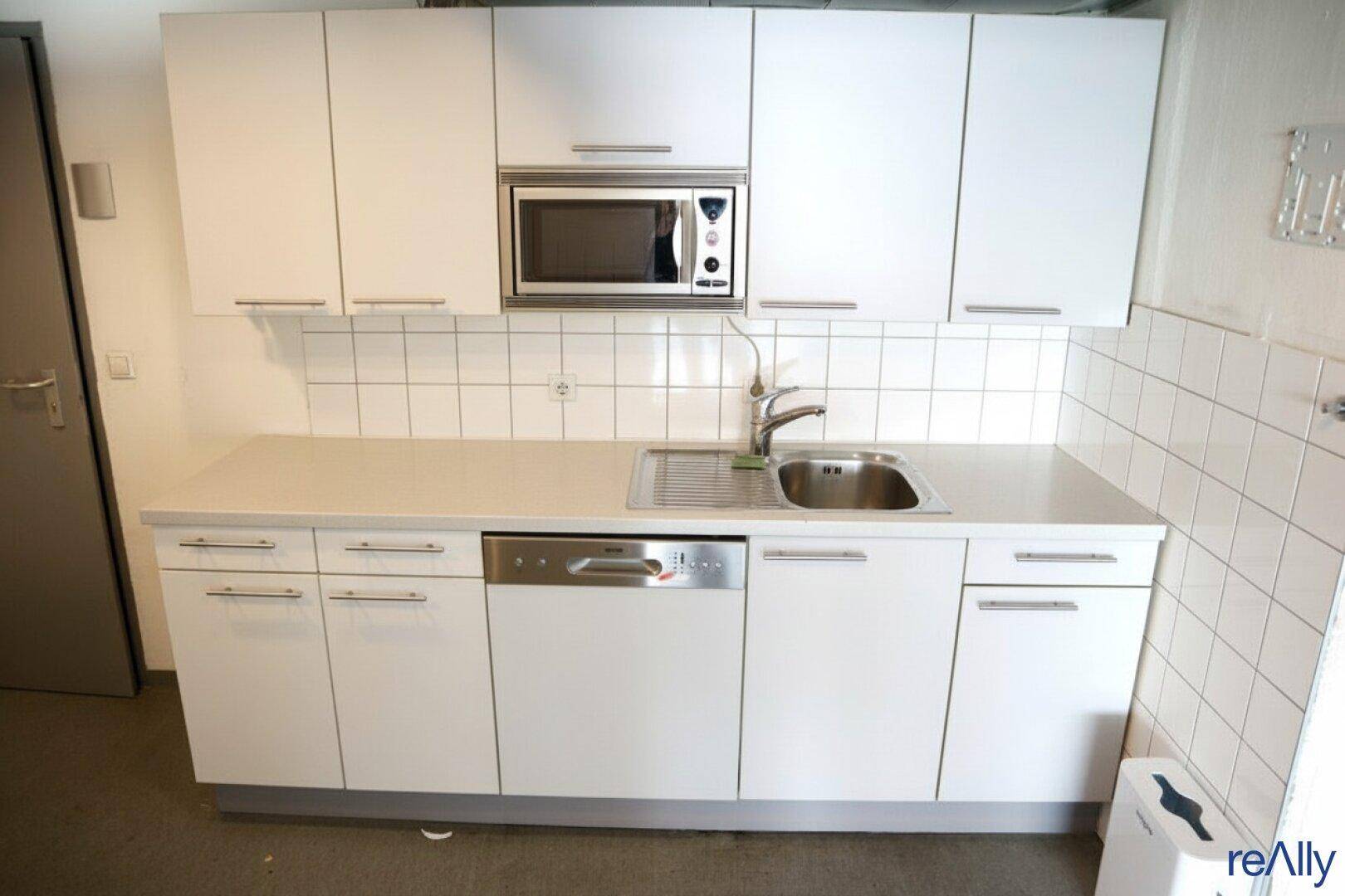 Kleine Einbauküche mit weißen Schränken, Spüle, Mikrowelle und Geschirrspüler.