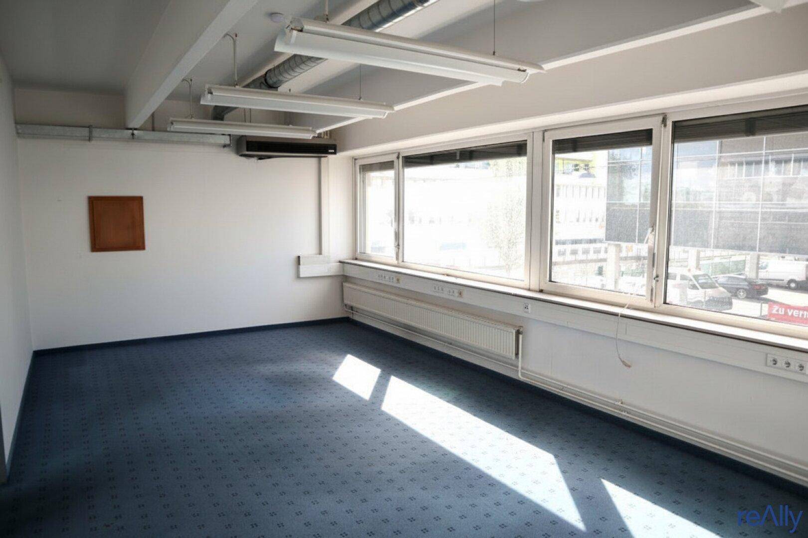 Helles Büro mit blauem Teppichboden, großen Fenstern und Blick auf die umliegende Stadtlandschaft.