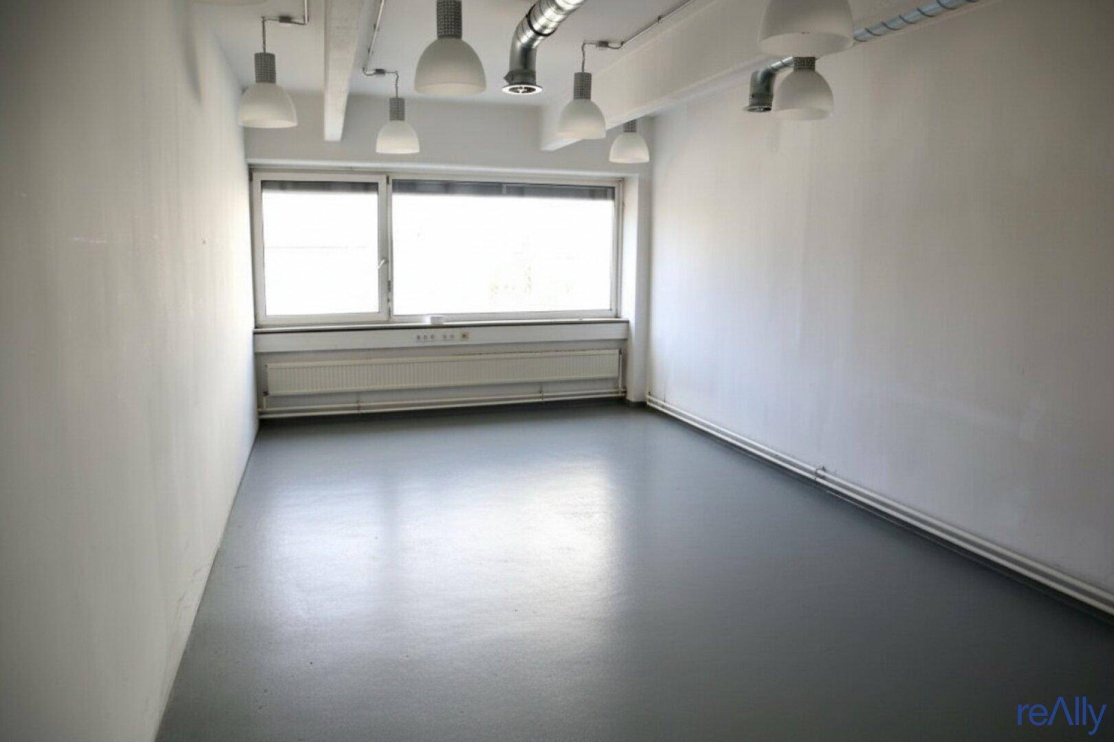 Büro mit grauem Boden, einem Fenster und sichtbaren Deckeninstallationen und Beleuchtung.