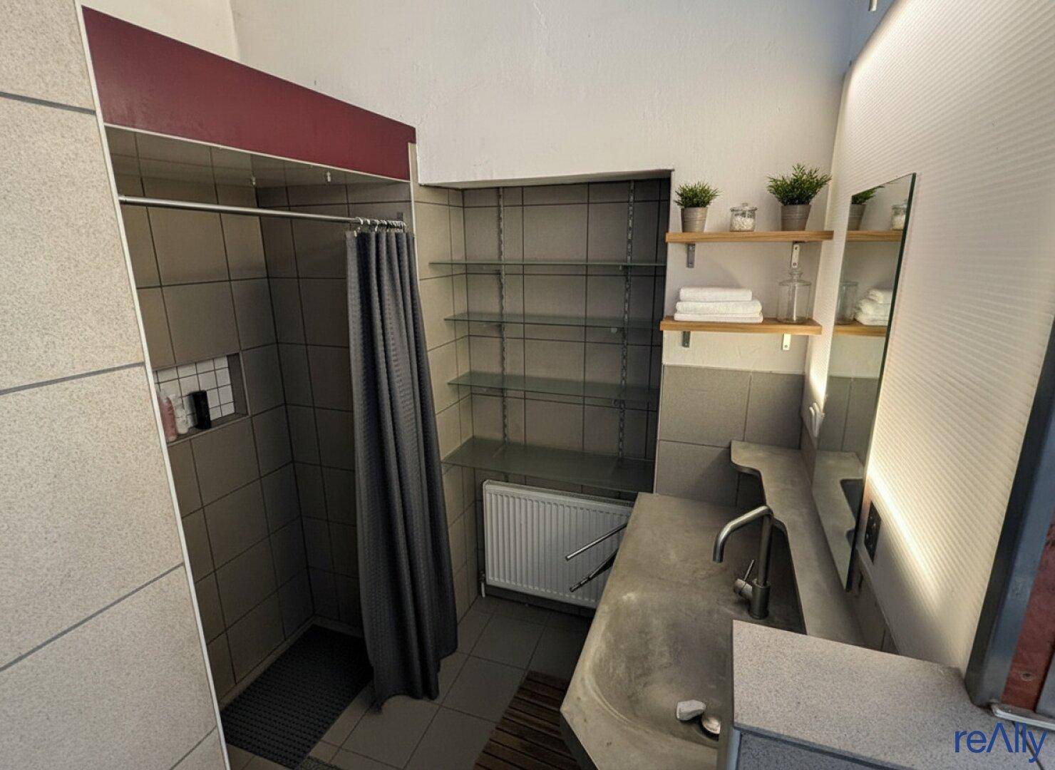 Badezimmer mit Duschbereich, Waschbecken und Regalen, in neutralen Grautönen gefliest.