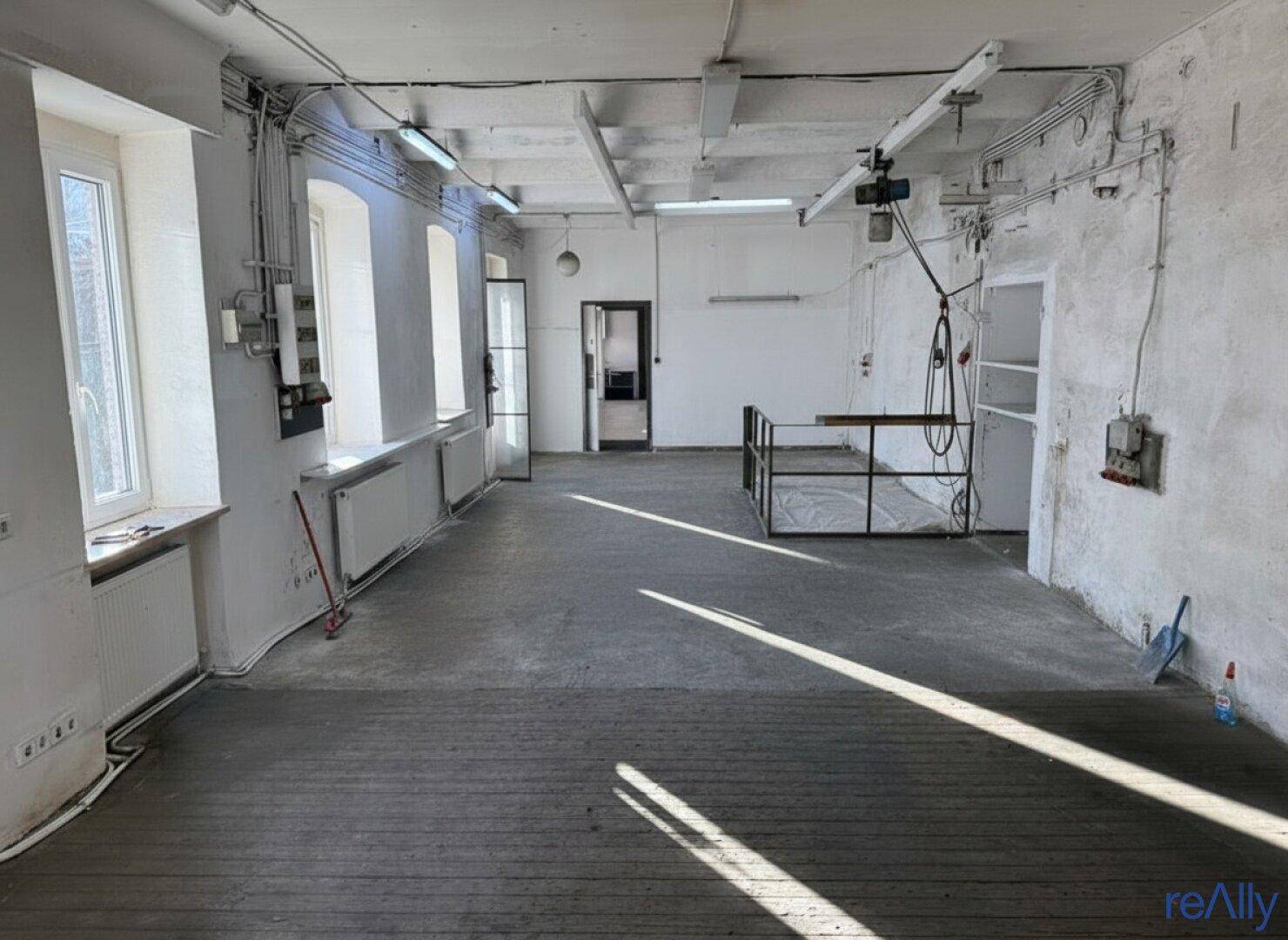 Großer, leerer Lagerraum mit Betonboden, vielen Fenstern und sichtbaren Installationen an Wänden und Decke.