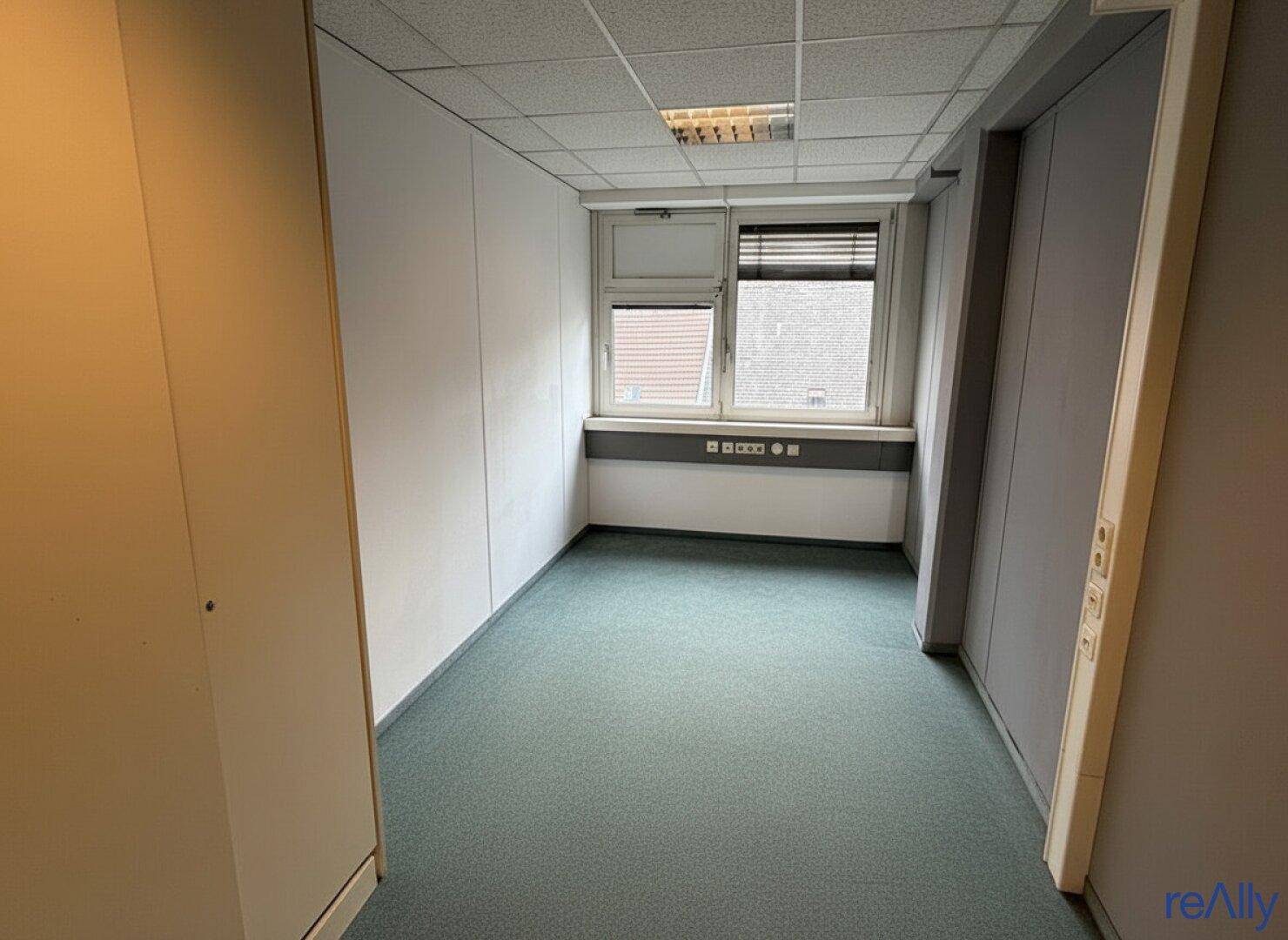 Kleineres Büro mit Teppichboden und einem Fenster, das einen Blick auf die umliegenden Gebäude bietet.