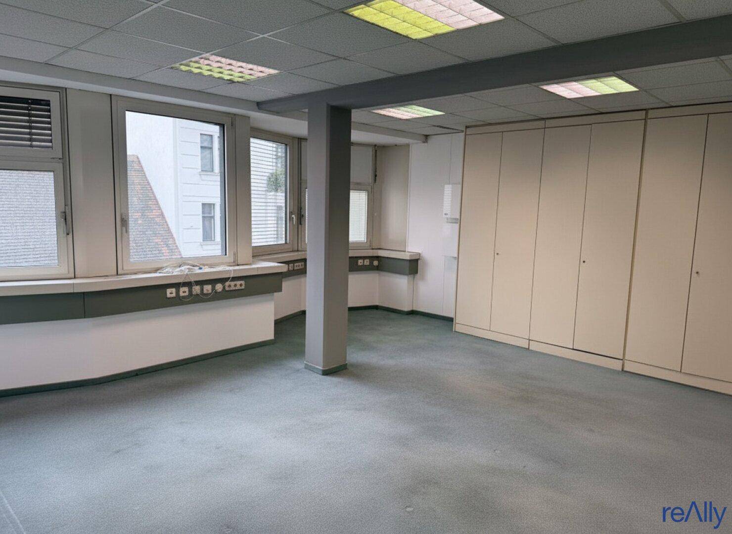 Großes Büro mit Teppichboden, vielen Fenstern und integrierten Schränken für effiziente Raumnutzung.