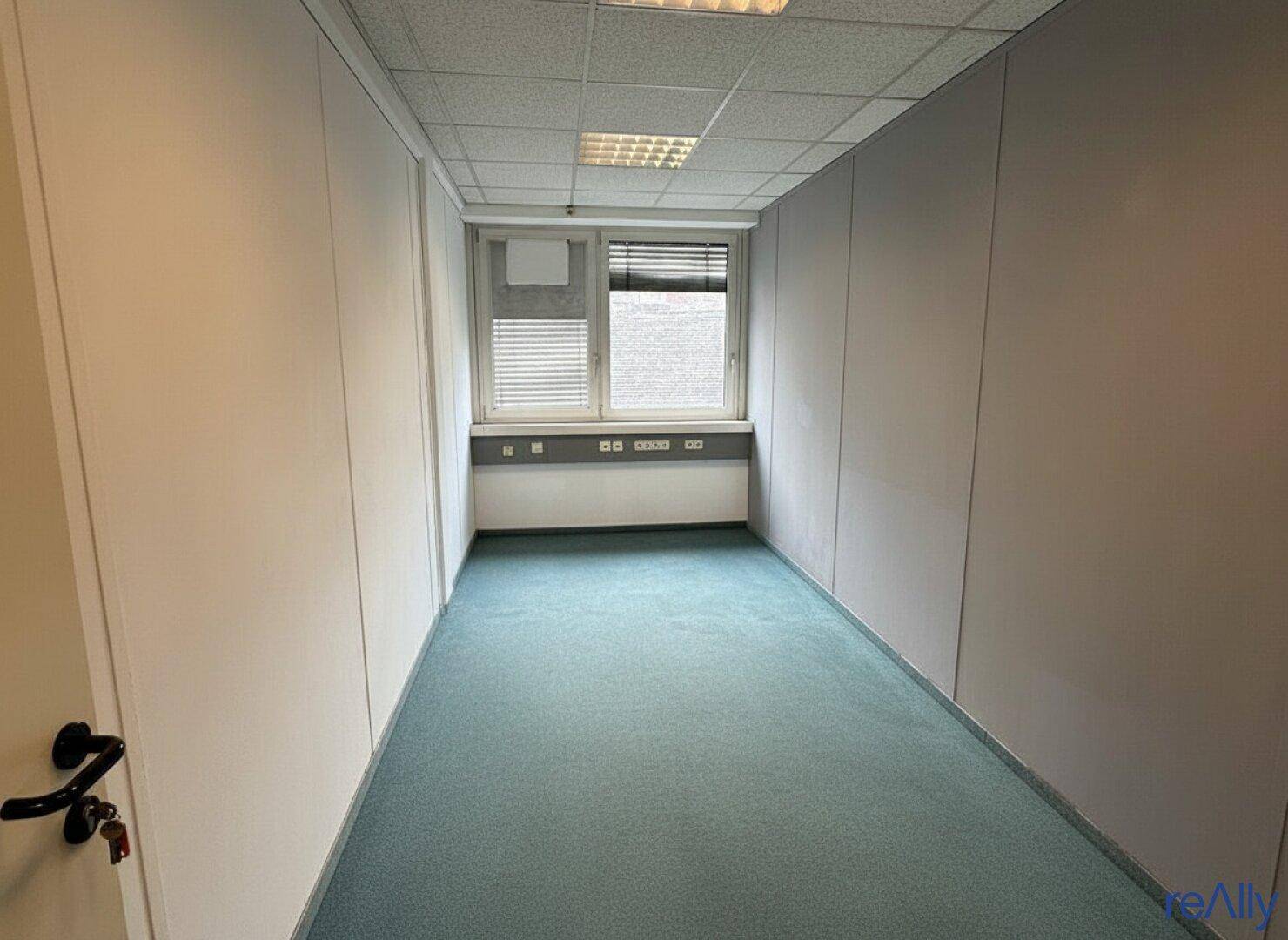 Schmaler Büroraum mit Teppichboden und einem Fenster, das ausreichend Tageslicht hereinlässt.