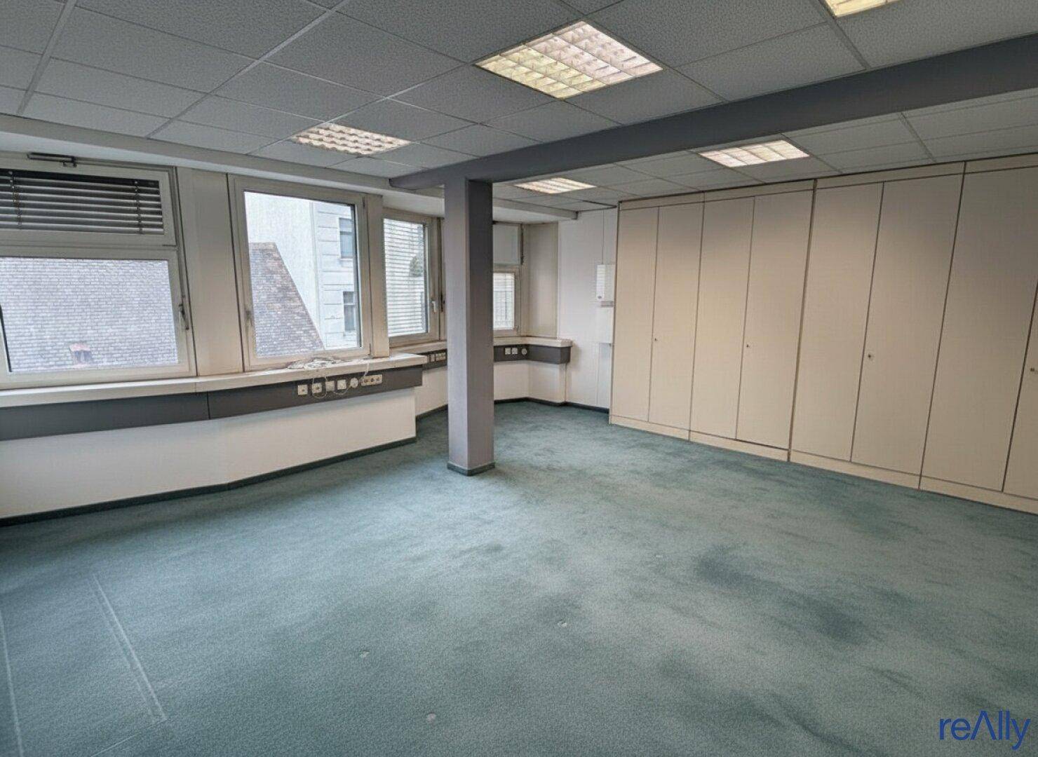 Geräumiges Büro mit Teppichboden, vielen Fenstern und integrierten Schränken entlang der Wände.