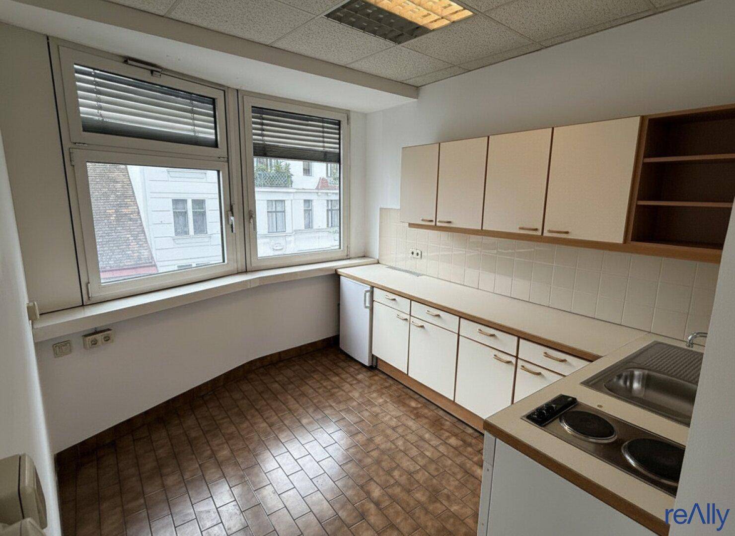 Kleine Küchenzeile mit weißen Schränken, Spüle und Kochplatten, ideal für eine Büroumgebung.