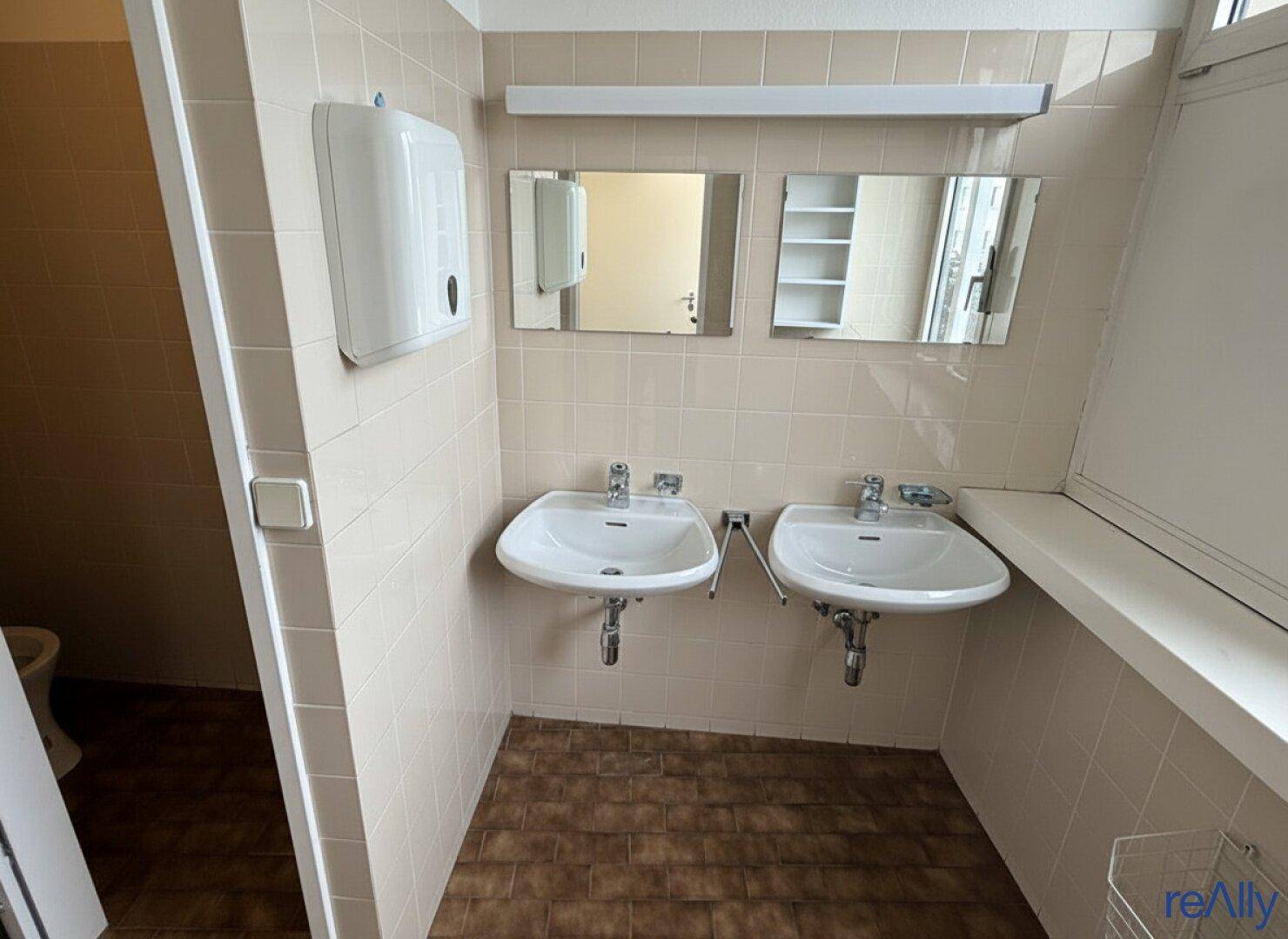 Sanitärraum mit zwei Waschbecken, Fliesenboden und einem Fenster für natürliche Belüftung.