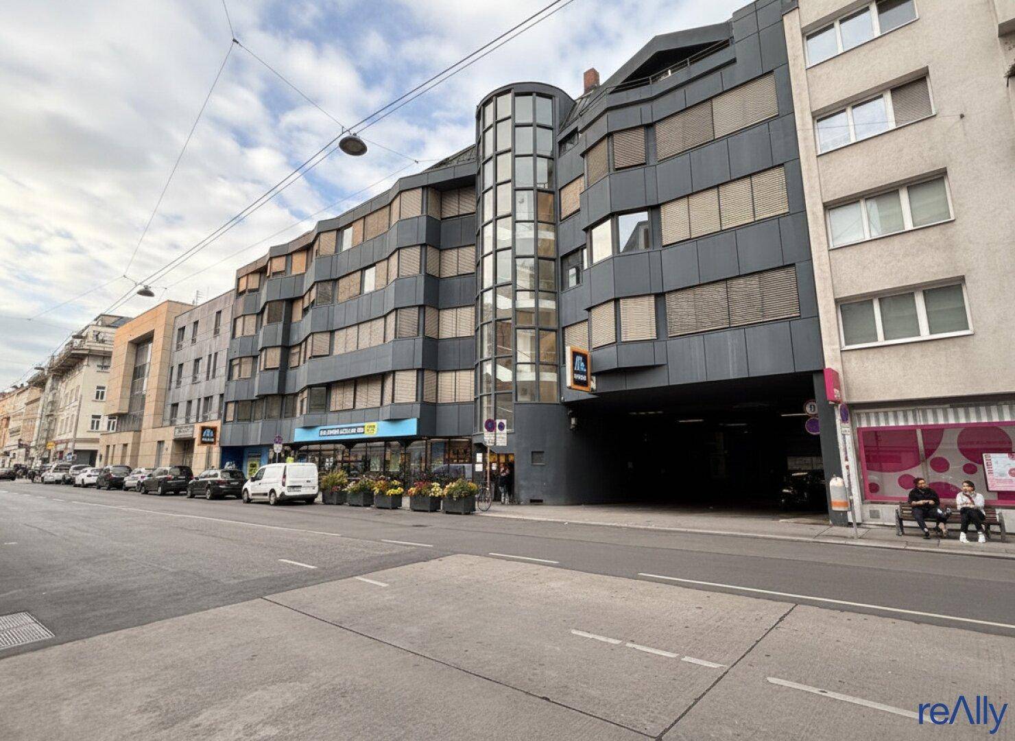 Breite Ansicht des modernen Gebäudes mit Geschäften im Erdgeschoss und Wohnungen darüber, an einer städtischen Straße.