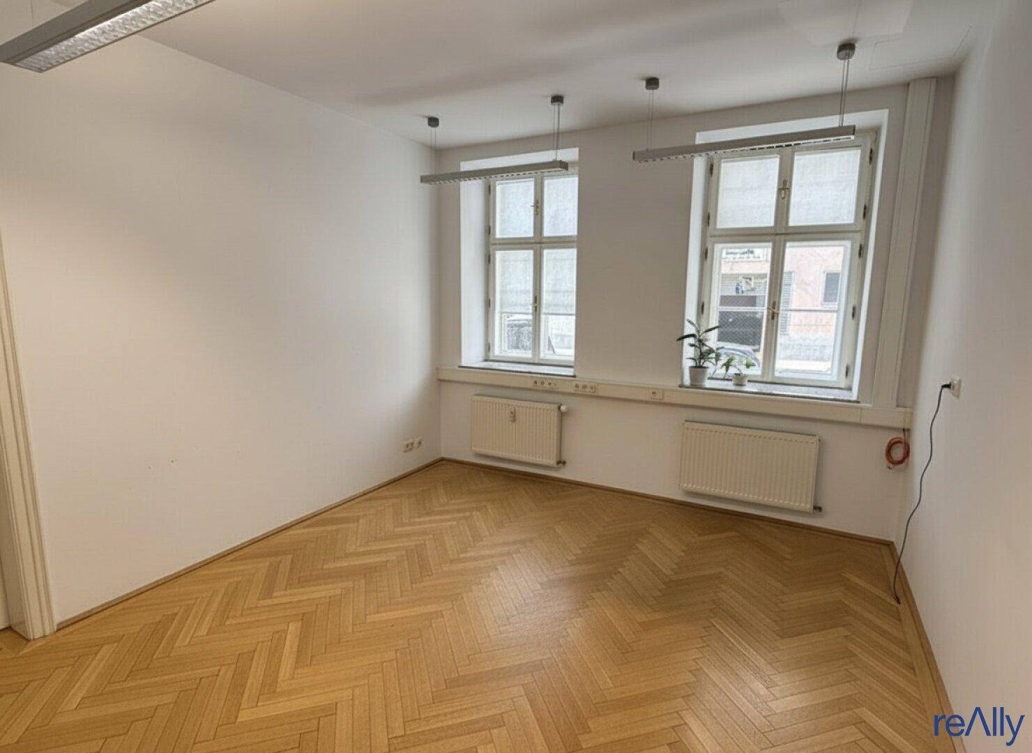Leeres Büro mit zwei großen Fenstern, Fischgrätparkett und viel natürlichem Licht.