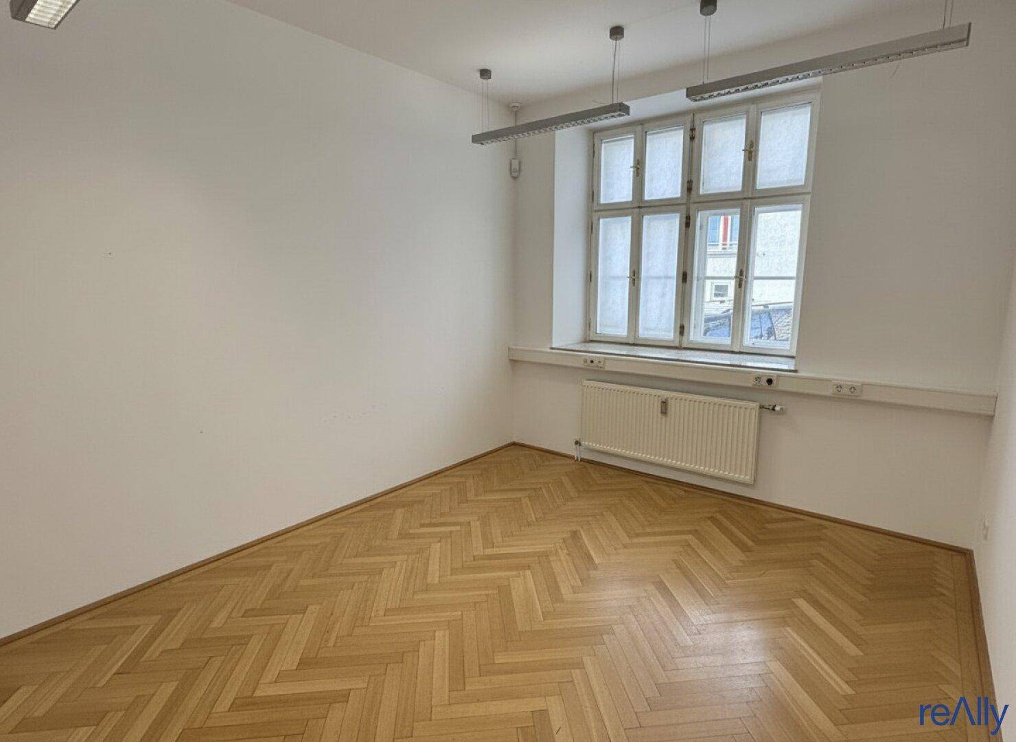 Helles Büro mit Fischgrätparkett und einem großen Fenster, das viel Tageslicht hereinlässt.