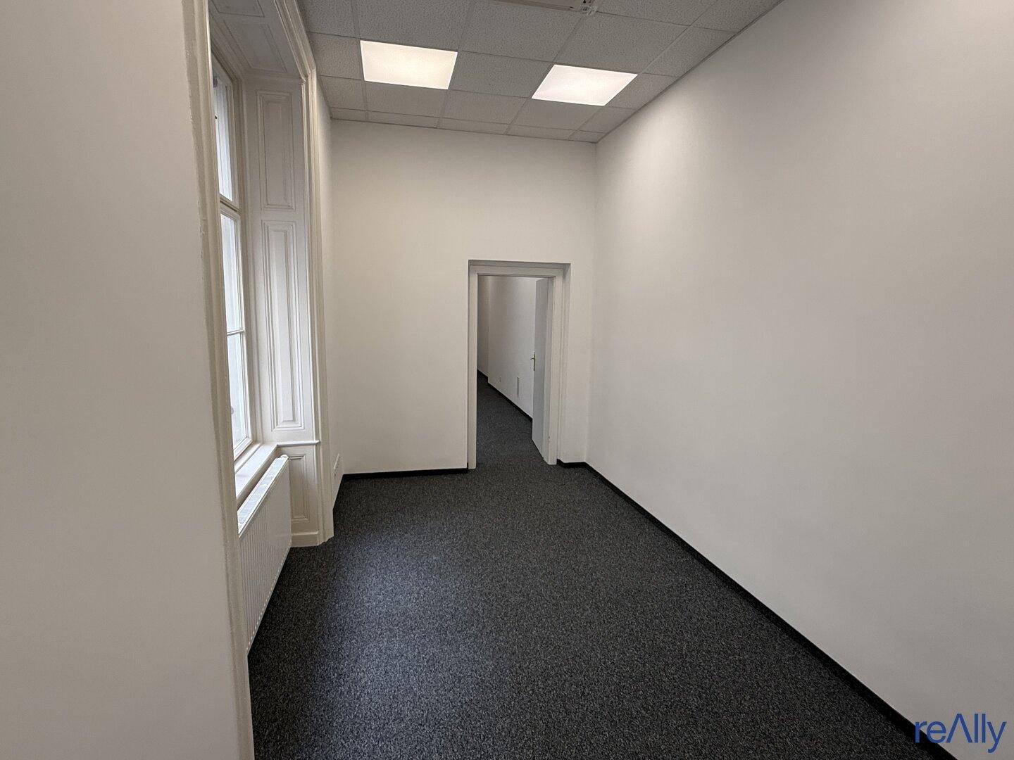 Schmaler Bürogang mit Teppichboden, weißen Wänden und einem Fenster, das Tageslicht hereinlässt.