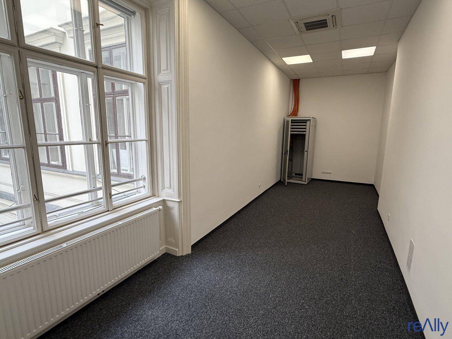Kleiner Büroraum mit Teppichboden, großem Fenster und einem Serverschrank, ideal für Technik.