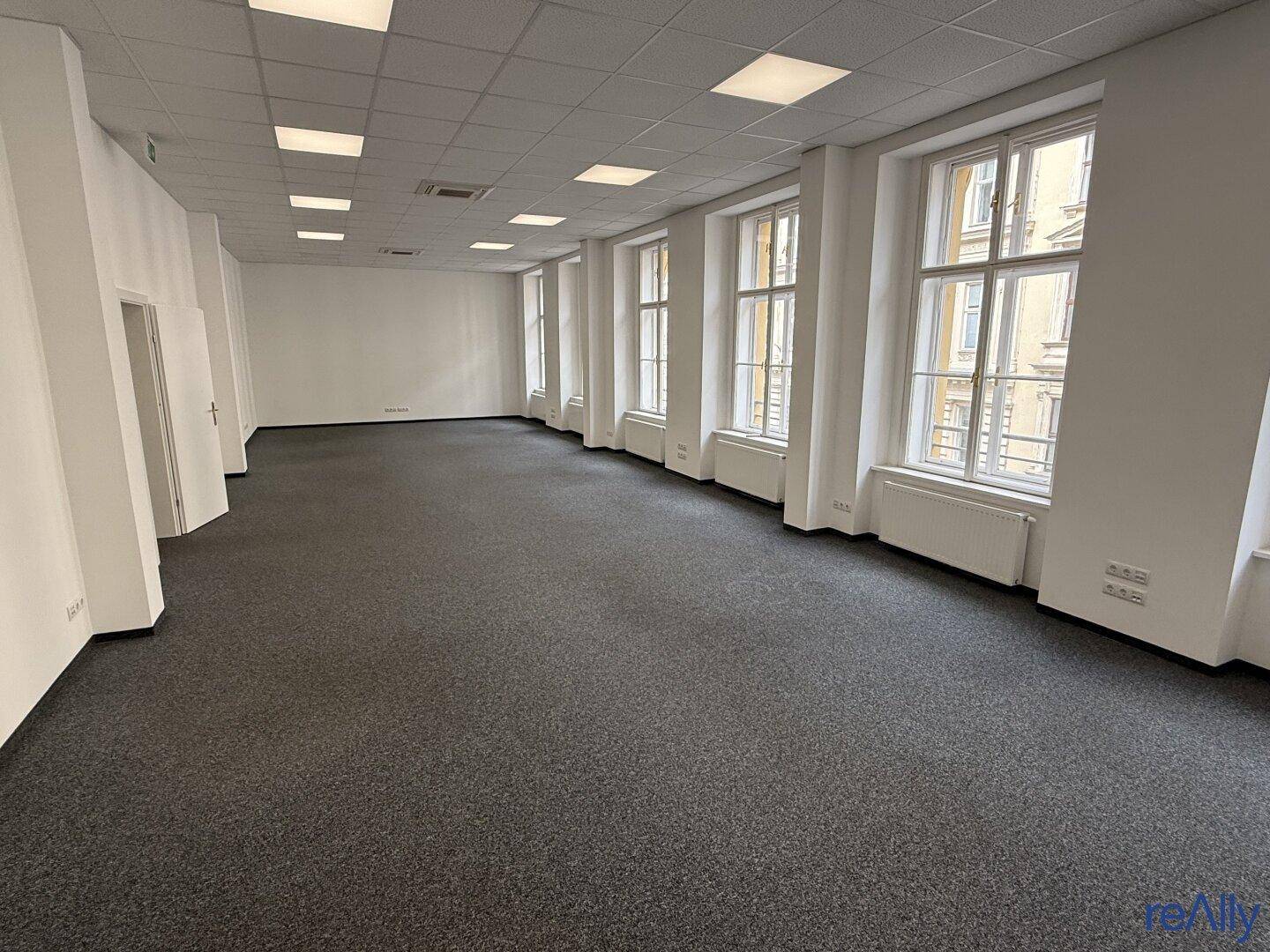 Geräumiger, leerer Büroraum mit Teppichboden und großen Fenstern, die viel Tageslicht hereinlassen.
