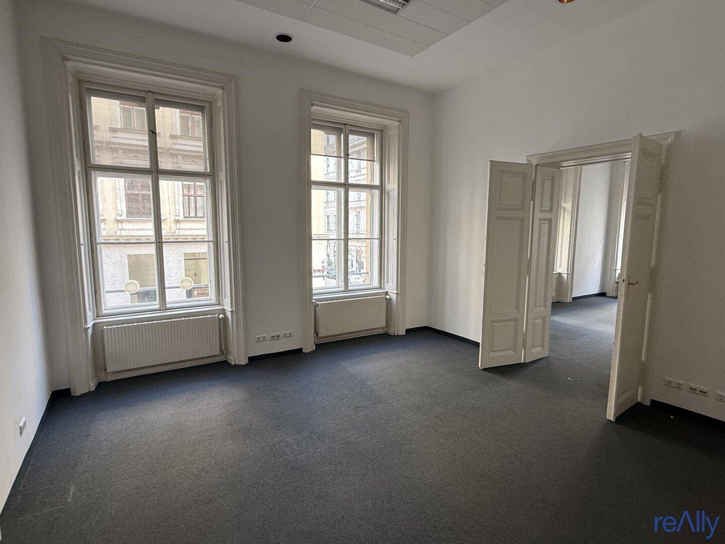 Heller, leerer Büroraum mit Teppichboden, zwei großen Fenstern und Zugang zu einem weiteren Zimmer.