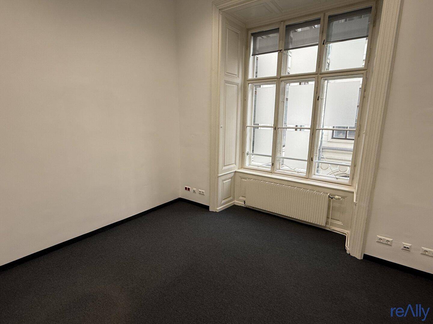 Leerer Büroraum mit Teppichboden, weißen Wänden und einem markanten Erkerfenster, das viel Licht spendet.