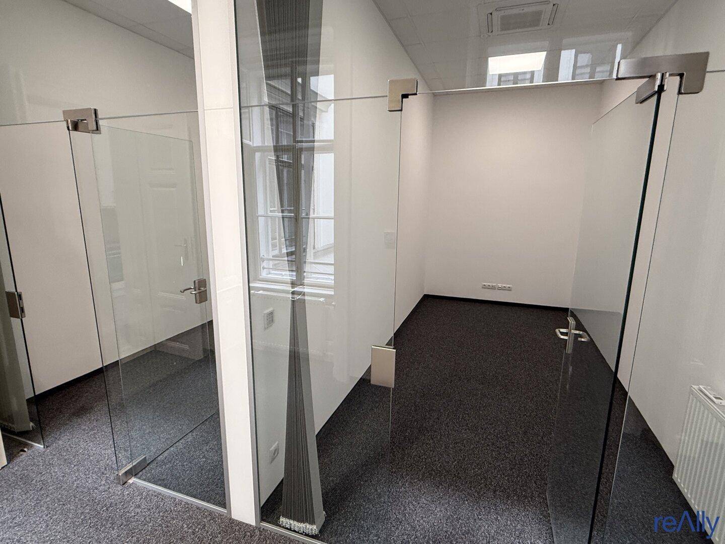 Zwei separate Büroräume mit Glaswänden und Teppichboden, die eine moderne und offene Arbeitsumgebung schaffen.