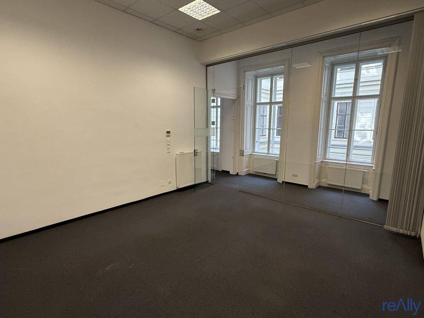 Helles Büro mit Teppichboden, großen Fenstern und einer modernen Glaswand als Raumteiler.