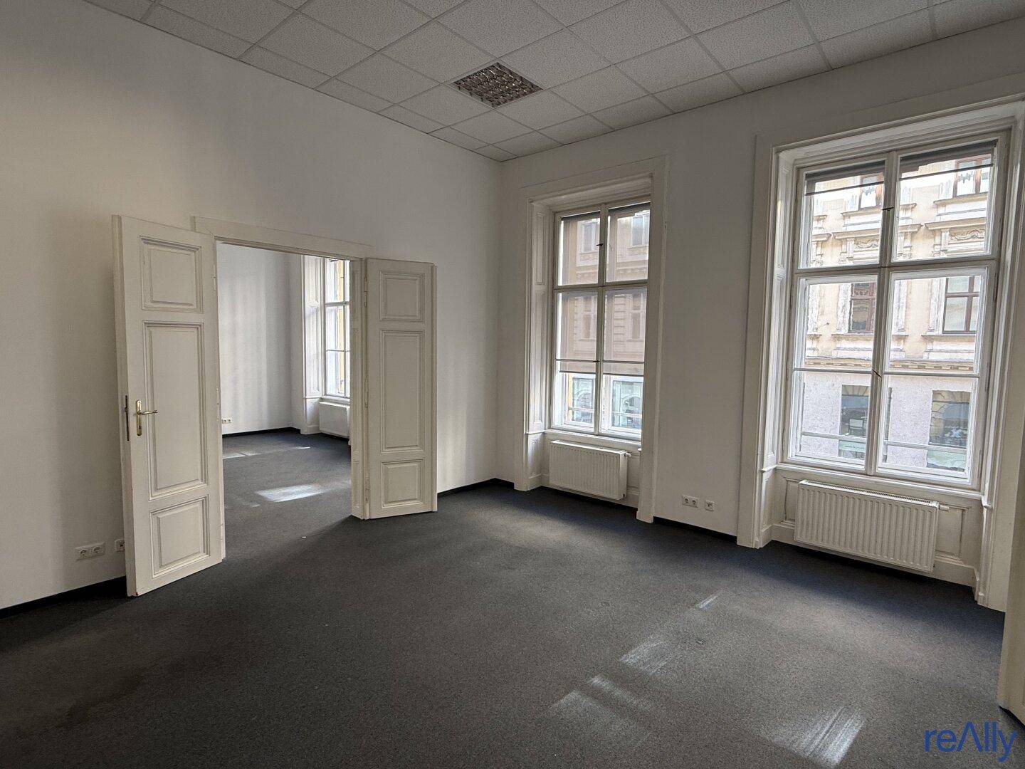 Geräumiges Büro mit Teppichboden, hohen Fenstern und weißen Doppeltüren zu einem Nebenraum.