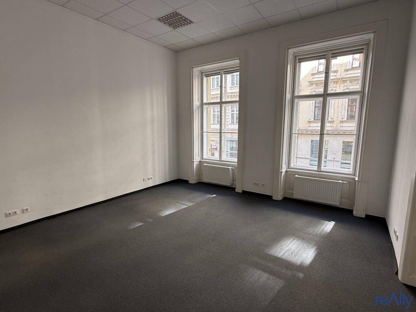 Heller Büroraum mit dunklem Teppichboden, hohen Decken und zwei großen Fenstern zur Straße.