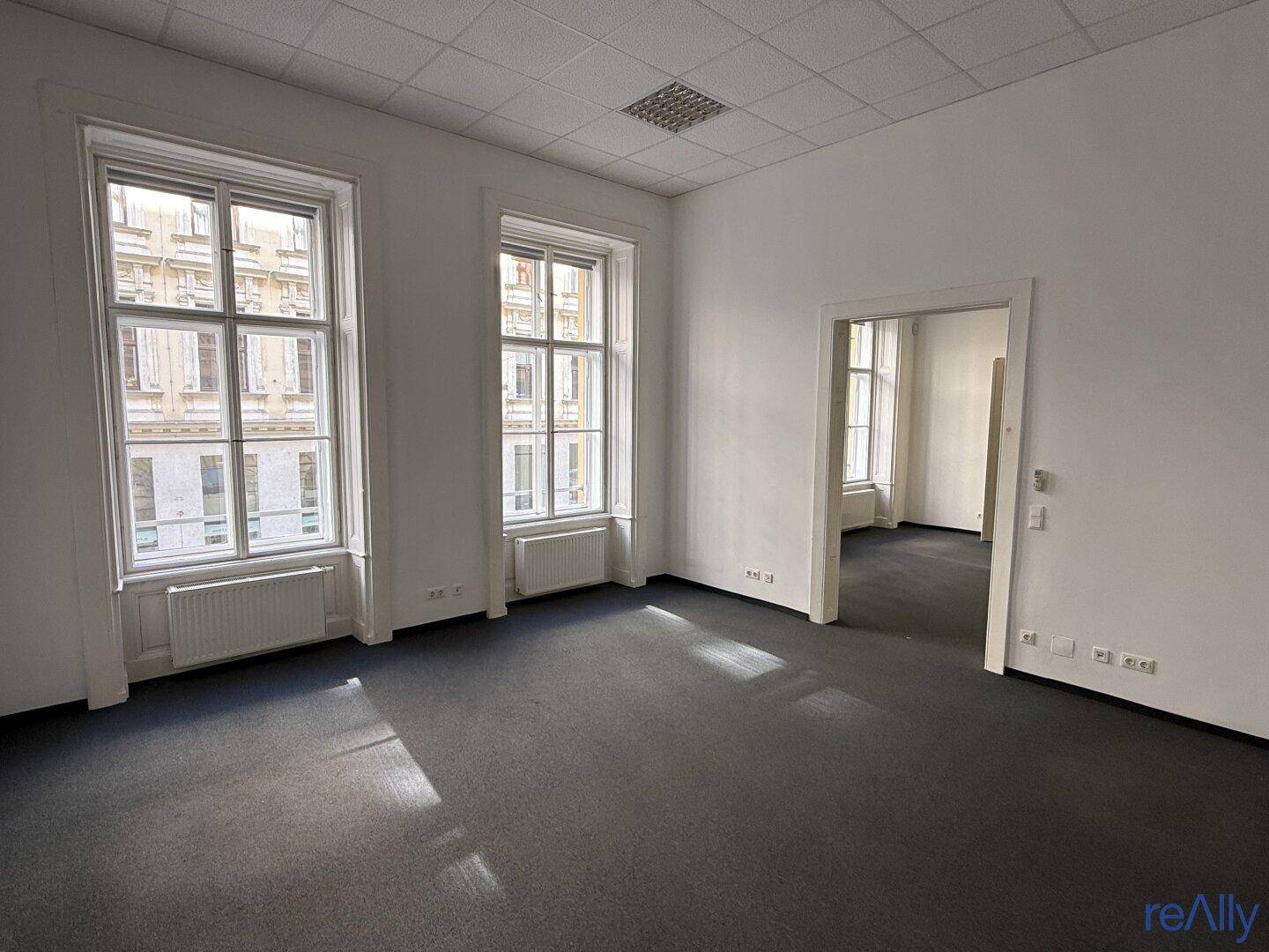 Leeres Büro mit Teppichboden, hohen Fenstern und einem Durchgang zu einem weiteren Raum.