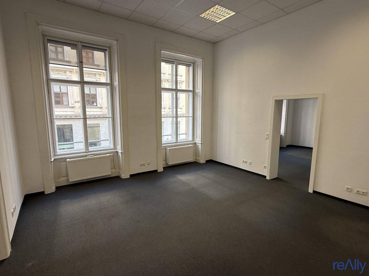 Leerer Büroraum mit Teppichboden, hohen Fenstern und einem offenen Durchgang zu einem weiteren Bereich.