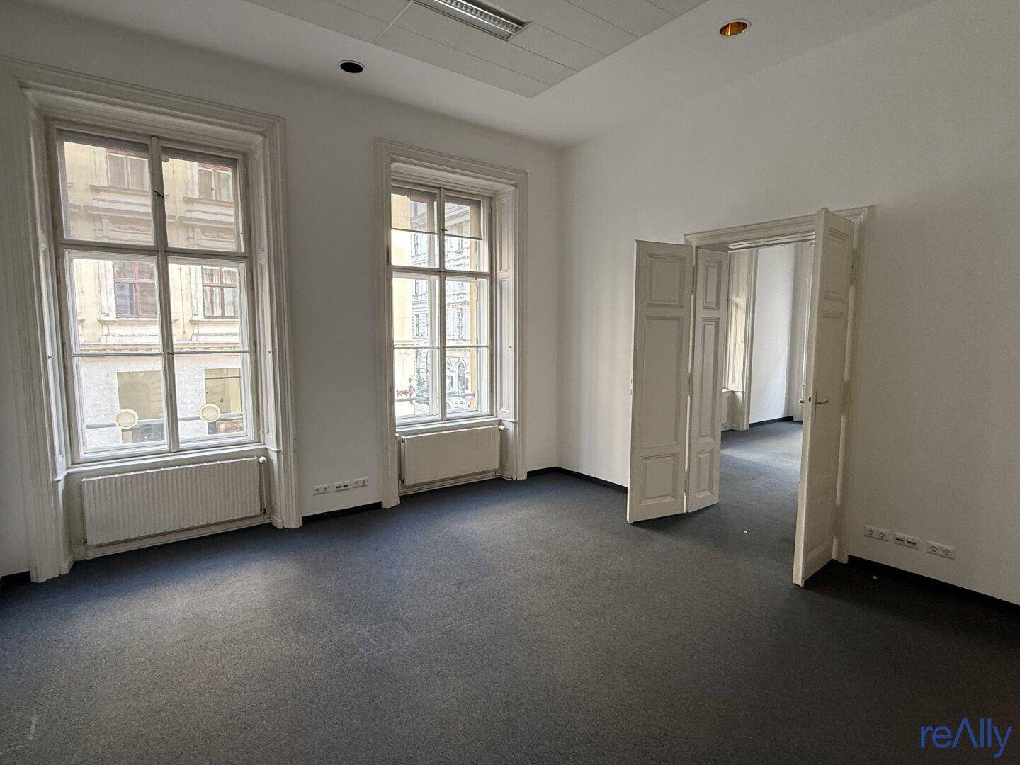 Heller Büroraum mit großen Fenstern, Teppichboden und Zugang zu weiteren Räumen.