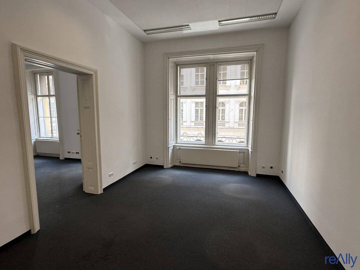Helles Eckbüro mit großen Fenstern, Teppichboden und Blick auf die umliegenden Gebäude.