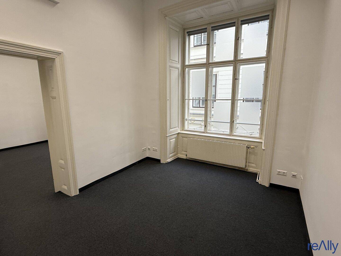 Heller Büroraum mit großen Fenstern, Teppichboden und Blick auf die gegenüberliegenden Gebäude.