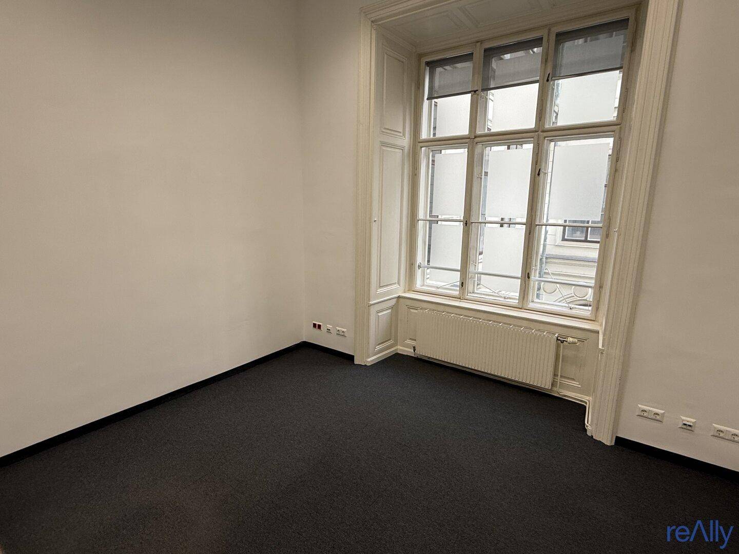 Ruhiger Büroraum mit Teppichboden, großen Fenstern und viel Tageslicht.