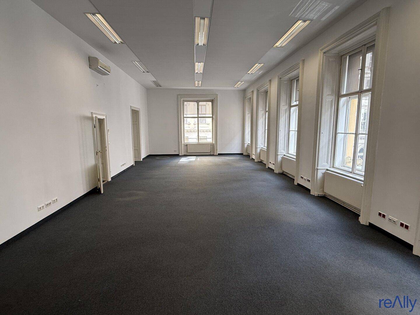 Geräumiges, leeres Büro mit Teppichboden, hohen Fenstern und Klimaanlage.