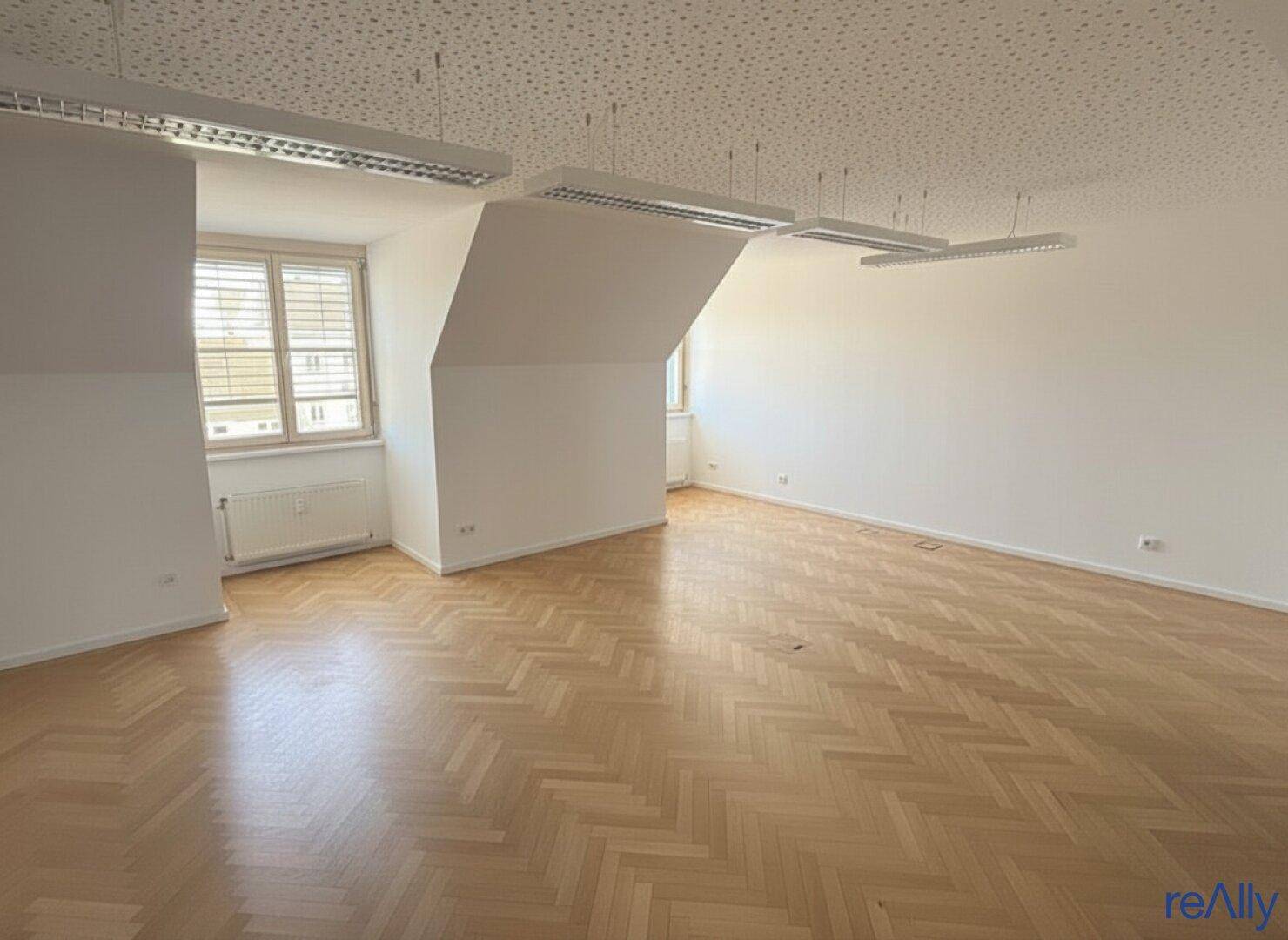 Heller Büroraum mit großen Fenstern, Fischgrätparkett und schrägen Wänden im Dachgeschoss.