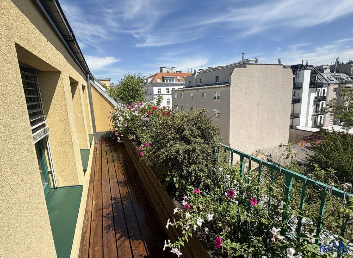 Langer Balkon mit Holzboden und üppiger Bepflanzung, bietet einen Blick auf die Stadt.
