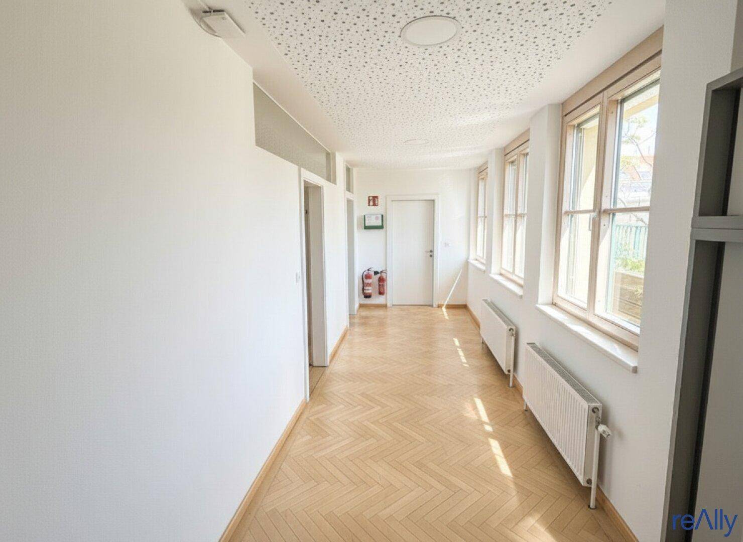 Heller Flur mit Fischgrätparkett, vielen Fenstern und Brandschutzvorrichtungen an der Wand.