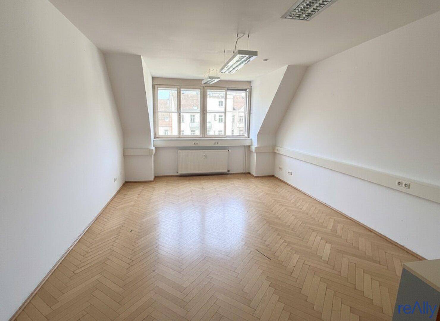 Leerer Büroraum mit Fischgrätparkett, Heizkörper unter dem Fenster und schrägen Wänden.