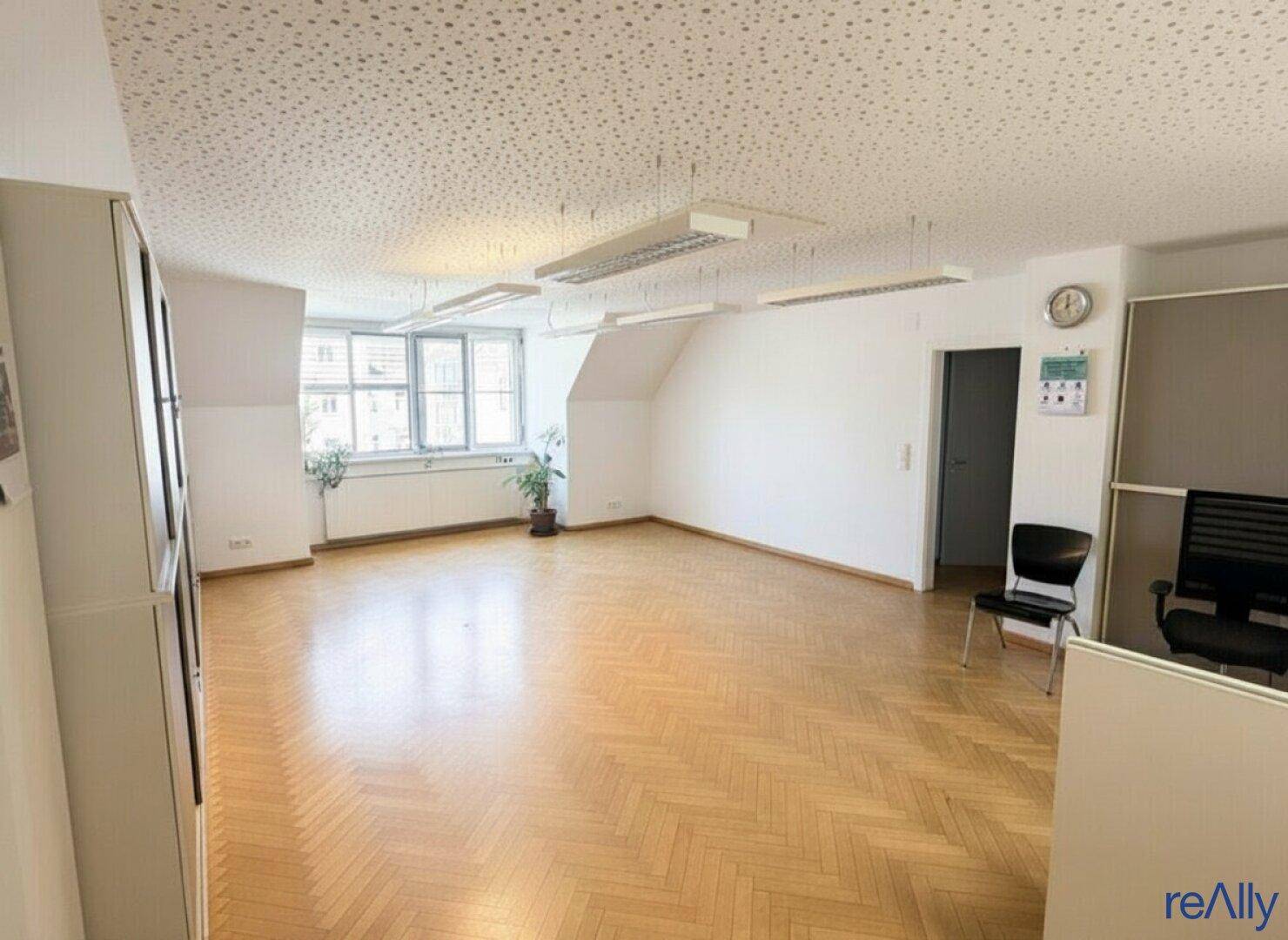 Großer Büroraum mit Fischgrätparkett, Fenstern und einer Uhr an der Wand.