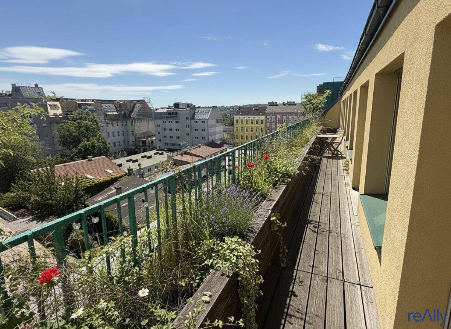 Langer Balkon mit Holzboden, üppiger Bepflanzung und weitem Blick über die Stadt.