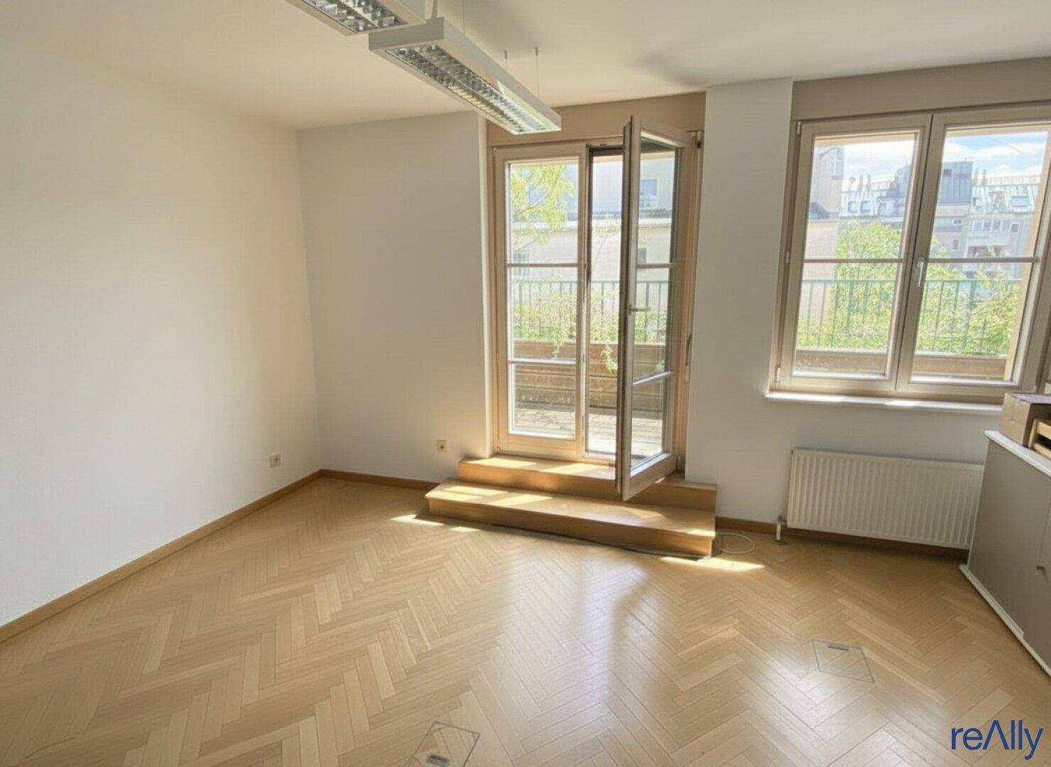 Heller Büroraum mit Zugang zu einem Balkon und großen Fenstern, ausgestattet mit Fischgrätparkett.