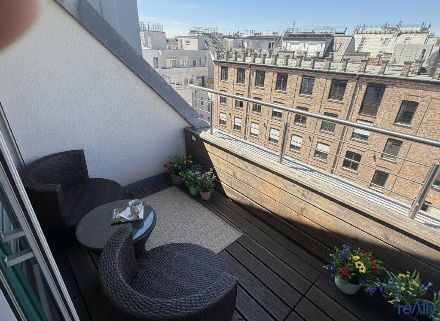 Kleiner Balkon mit Holzboden, Sitzgelegenheiten und Blick auf die umliegenden Gebäude.