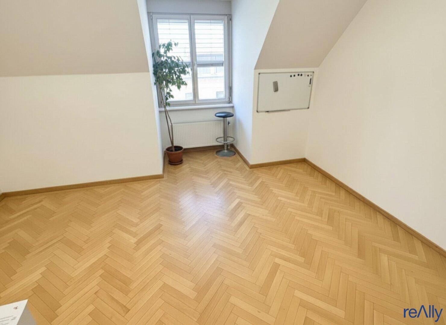 Heller Büroraum mit Fischgrätparkett, Fenster und einem kleinen Schrank an der Wand.