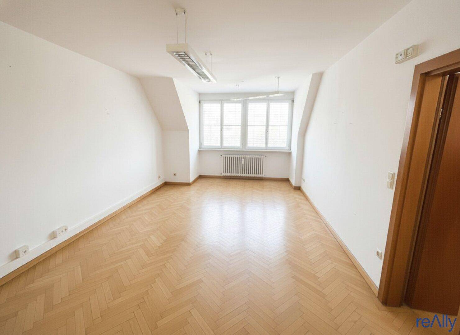 Geräumiger Büroraum mit Fischgrätparkett, großen Fenstern und einer Holztür.