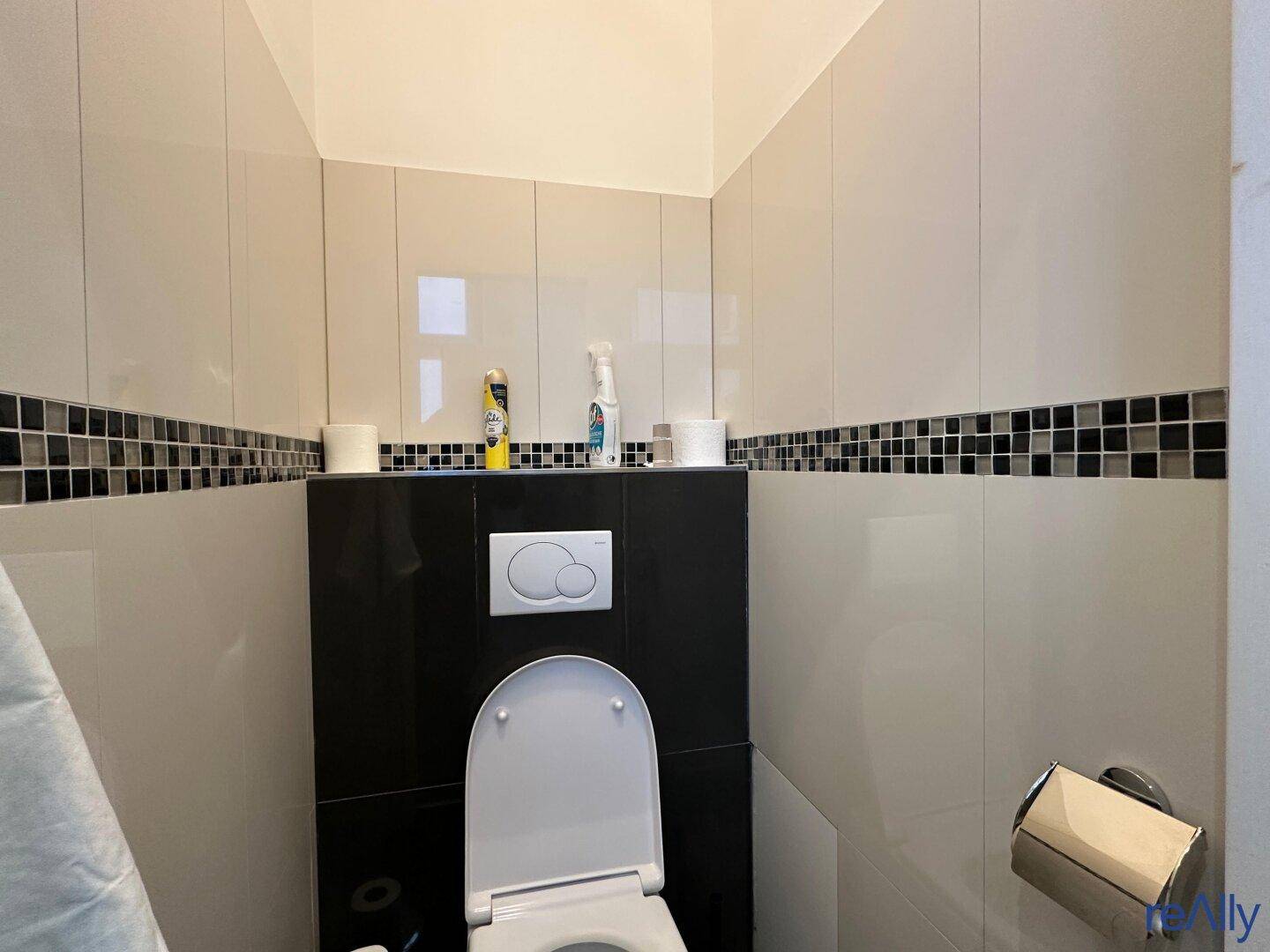 Modern gefliestes WC mit Wand-WC und dunkler Akzentleiste.