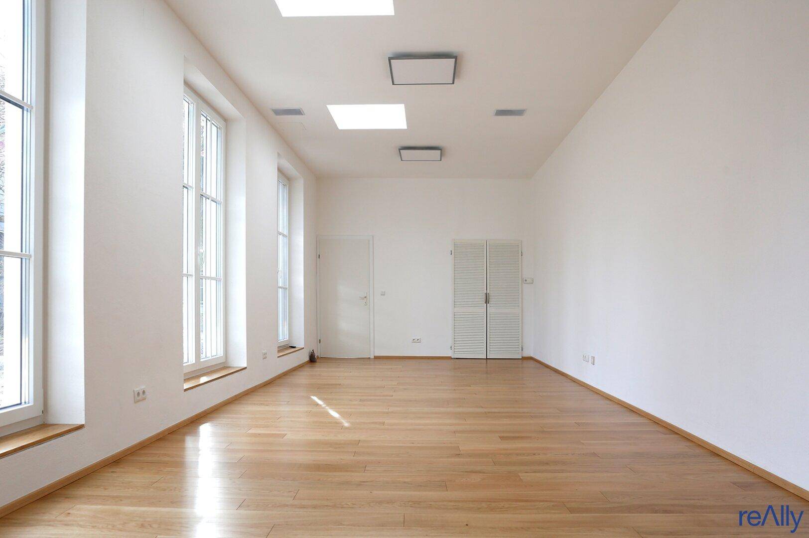 Heller, leerer Raum mit hohen Fenstern und glänzendem Holzparkettboden.