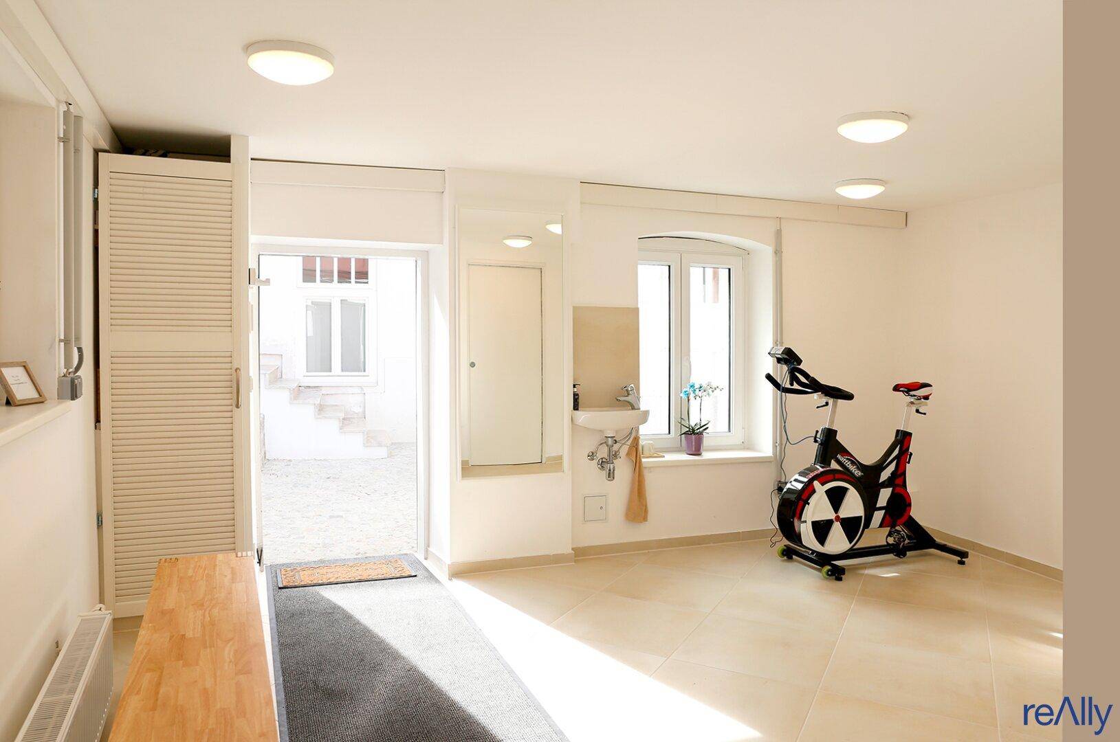 Fitnessraum mit Trainingsrad, Fliesenboden und Zugang zu einem Außenbereich.
